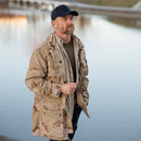 Man wearing navy blue military style baseball cap and beige camouflage jacket by the water at sunset