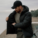Man wearing black Pentagon Omega down jacket with hood, showing the inside, standing outdoors near water.
