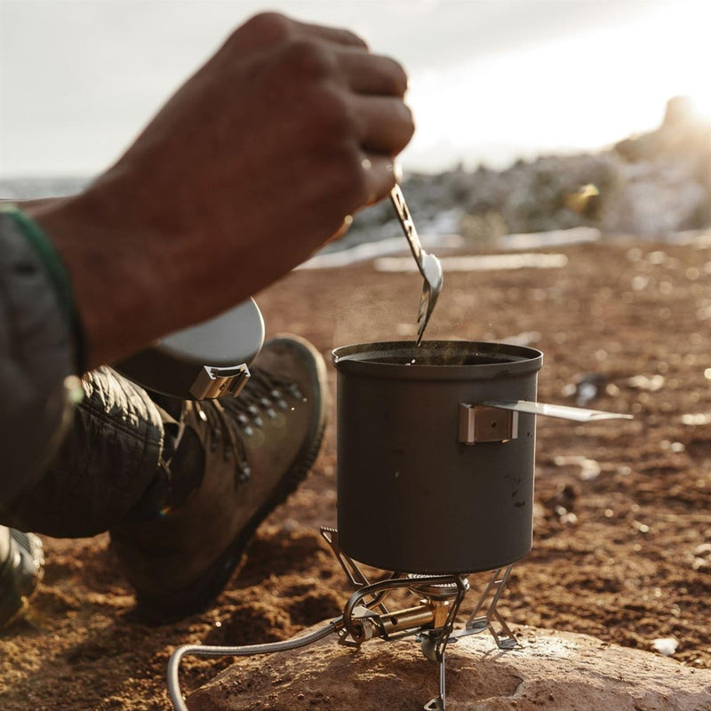 Person stirring food in a camping pot on a Primus Express Spider II tripod stove outdoors at sunset