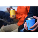 Person holding blue Primus Trailbreak insulated vacuum jug outdoors with another person in the background holding a yellow jug